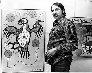 A black-and-white photograph of a young Norval Morrisseau, taken in 1977, standing in front of a canvas of his own artwork.